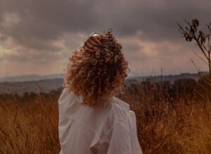 Back view of unrecognizable female touching curly hair while contemplating cloudy sky and mounts with faded grass in evening