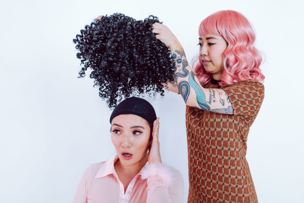 A hair stylist adjusts a curly wig on an adult woman indoors, showcasing fashion and beauty.